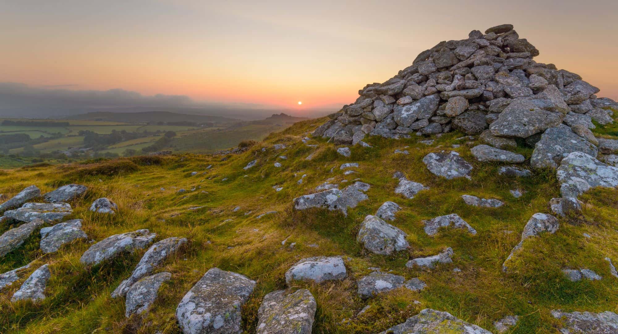 A rocky hilltop scene in Dartmoor at sunset, with a cairn of stacked stones in the foreground, set against a warm orange sky. The landscape features grassy terrain interspersed with rocks, rolling into a lush, green valley below with distant hills obscured by mist.