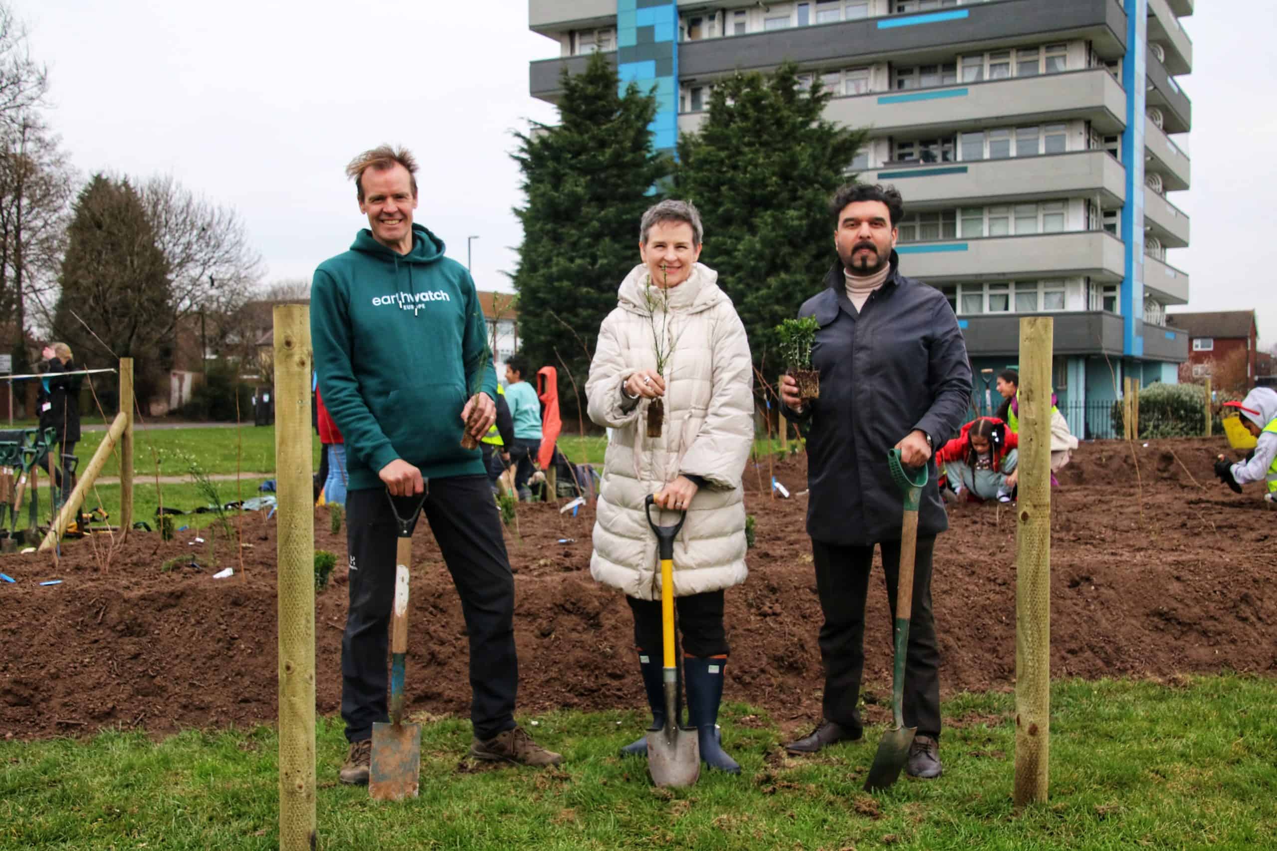 Earthwatch CEO, Harry Barton, Mary Creagh, MP, and Councillor Khan at the Bell Green Road Tiny Forest planting day in Coventry. There is a high rise building in the background. They are holding small tree saplings and smile into the camera.